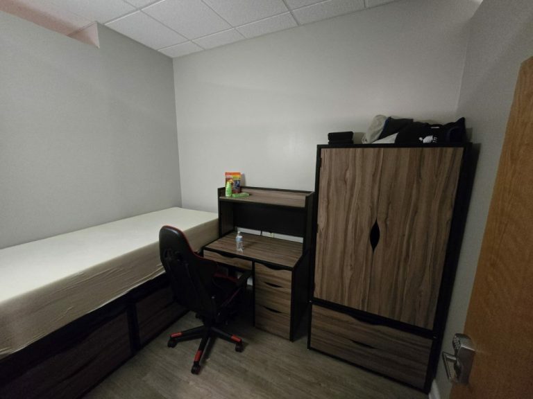 A small room with a single bed, a wooden desk with a chair, and a closet. The desk has a few items, including a water bottle and snacks. The walls are plain, and there's a light wooden floor.