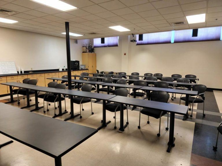 An empty classroom with black tables and chairs arranged in rows. The room has a whiteboard on the left, cabinets at the back, and a projector mounted on the ceiling. The windows are covered with blinds.