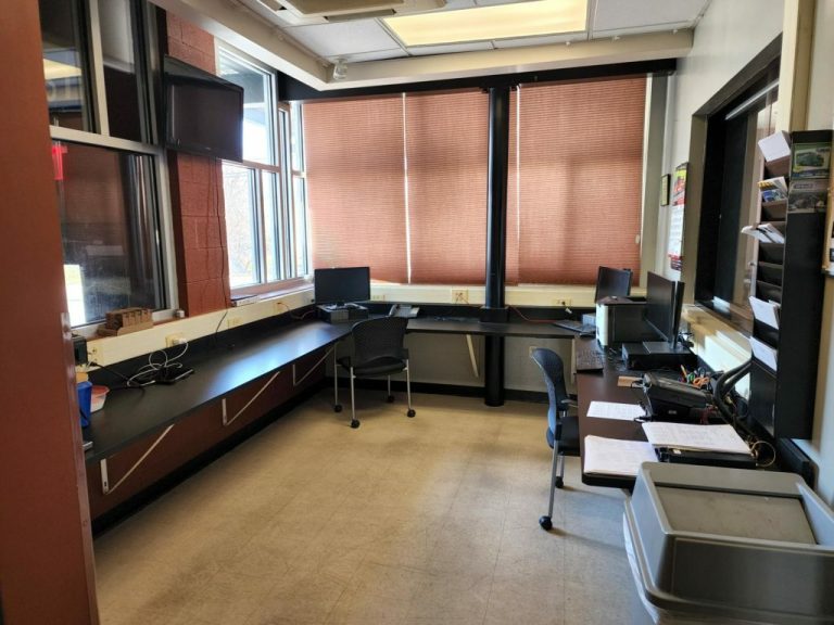 A small office space with large windows, brown blinds, and several workstations. There are two black chairs, two monitors, scattered papers, and shelves with miscellaneous items. The floor is light-colored tile.