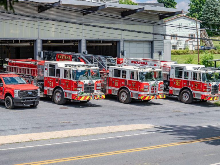 A row of fire trucks and an emergency vehicle are parked in front of a fire station. The trucks are red with "SWATARA" written on them, and the station has open garage doors. The scene is sunny with some trees in the background.