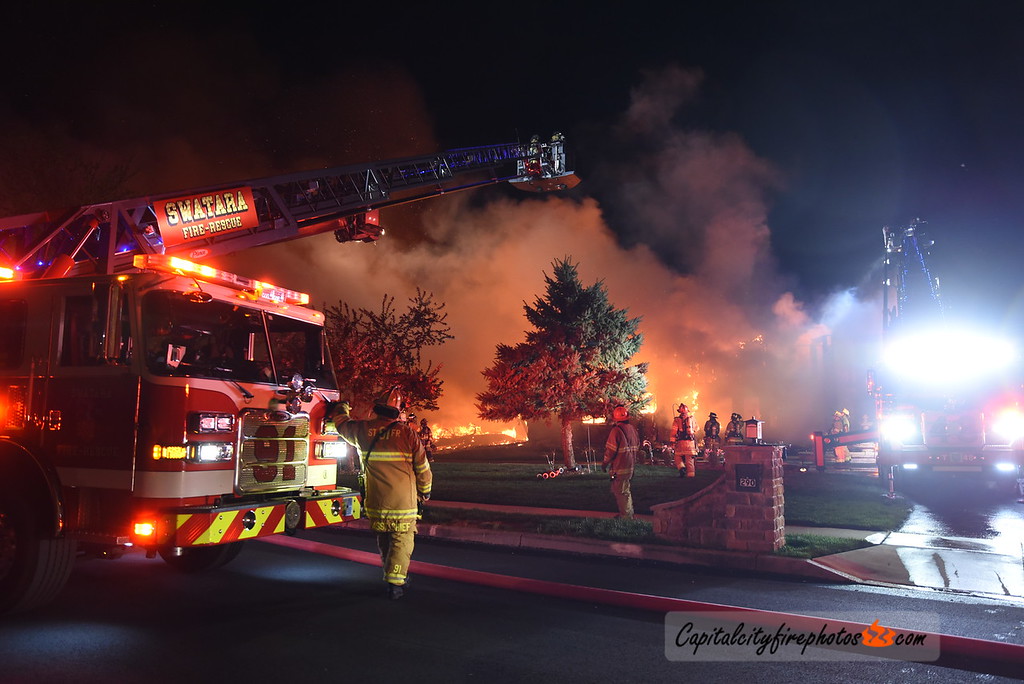 Firefighters respond to a large fire at night, with fire trucks and a ladder in action. Bright flames and smoke are visible, illuminating the scene. Emergency crews work to control the situation.