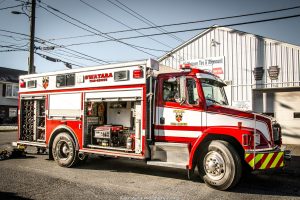 A red and white fire rescue truck is parked on a sunny street. The truck's compartments are open, revealing equipment. Buildings and power lines are visible in the background, with shadows cast on the pavement.