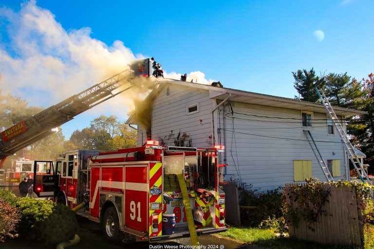 Firefighters on a ladder truck battle a house fire with smoke billowing from the roof. Red fire trucks with flashing lights surround the white two-story building. Sunny day with clear skies and trees in the background.