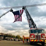 A large American flag is suspended between two extended fire truck ladders against a cloudy sky. The fire truck is parked on a wide road, with people and motorcycles visible in the background.