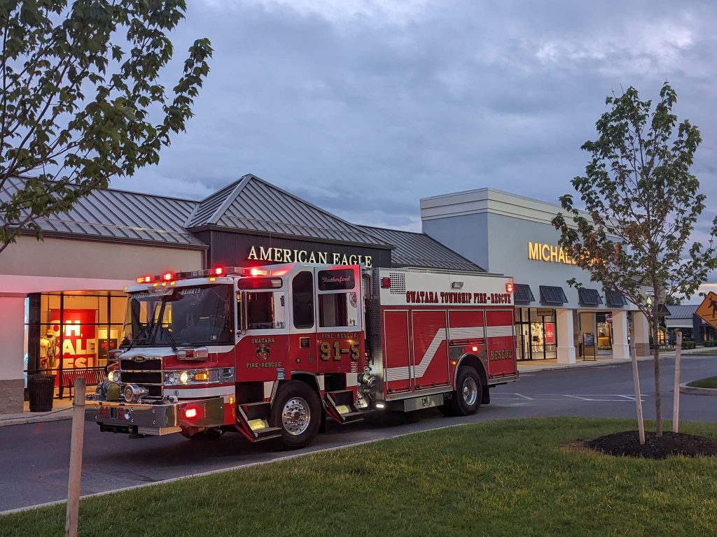 A red and white fire truck is parked in front of shops, including American Eagle and Michael Kors. The sky is overcast, and there are small trees lining the pavement. A "sale" sign is visible inside the American Eagle store.