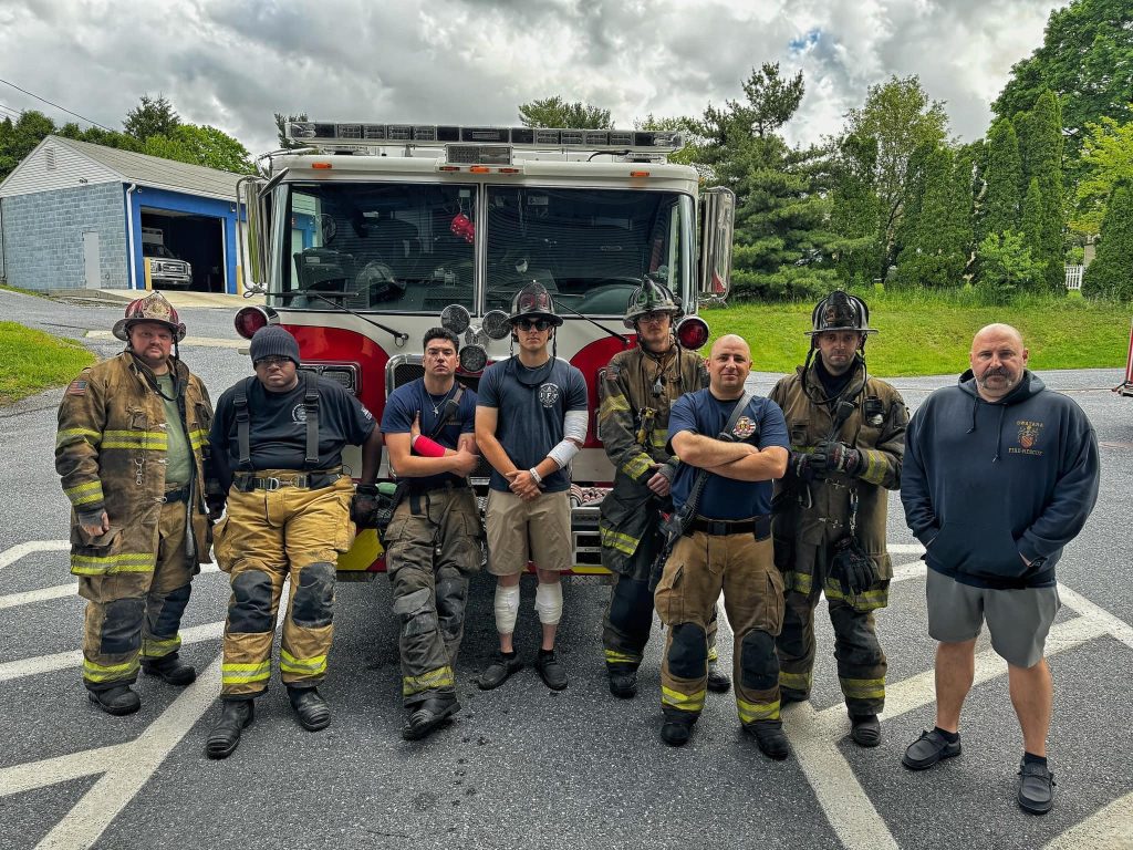 A group of firefighters stands in front of a fire truck, with some wearing full gear and helmets. They are outdoors on a paved surface with trees and a building in the background. The sky is cloudy.