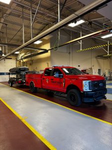 A red emergency response truck labeled "Swatara Fire Rescue" is parked inside a garage. The truck is towing a trailer with a rescue boat. The interior features yellow and red painted lane markings and overhead equipment.