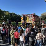 A large group of children stands outside, listening to a firefighter beside a fire truck. The truck's doors are open, revealing equipment. Adults are also present. The sky is clear and blue, with green trees in the background.