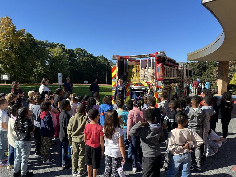 A large group of children stands outside, listening to a firefighter beside a fire truck. The truck's doors are open, revealing equipment. Adults are also present. The sky is clear and blue, with green trees in the background.