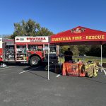 A Swatara Fire-Rescue truck is parked in a lot. A red tent with the same branding has a table underneath displaying fire safety materials. A person is standing behind the table. Trees and a clear sky are in the background.