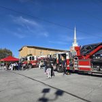 Fire trucks parked near a church with a prominent steeple under a clear blue sky. People, including children, gather around a red canopy tent and the trucks. Trees and a building are visible in the background.