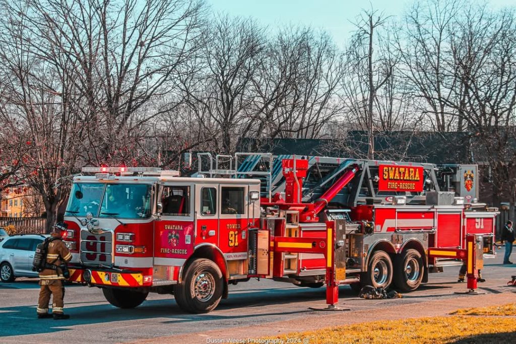 A fire truck with Swatara Fire Rescue branding is parked on a street. A firefighter wearing a helmet and uniform stands near the truck. The background features leafless trees and a clear sky.