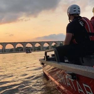 A person wearing a helmet and life jacket sits on a boat at sunset. The water reflects the warm hues of the sky. An arched bridge stretches across the background.