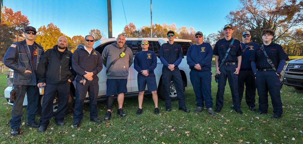A group of ten people, some in dark uniforms and others in casual clothing, stand in front of a vehicle on a grassy area. Trees with autumn leaves are in the background. They appear to be firefighters or emergency personnel.