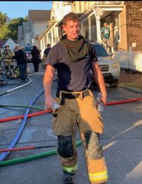 A firefighter in gear stands on a sunlit street holding a water bottle, with hoses and emergency personnel visible in the background near residential houses.