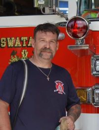 A man with a mustache, wearing a navy blue shirt with a fire department emblem, stands in front of a red fire truck. He has a shoulder bag and a necklace with a cross.