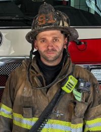 A firefighter in full gear and helmet stands in front of a fire truck, looking at the camera. His uniform is dirty, indicating he has been working at a scene.