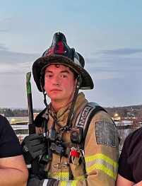 A firefighter in full gear, including a helmet and reflective jacket, stands outdoors at dusk holding an axe, with buildings and a cloudy sky in the background.