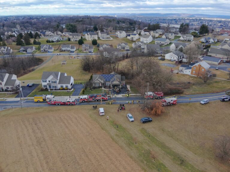 Aerial view of a residential neighborhood with several fire trucks and emergency vehicles parked along a road near a house, as firefighters and people gather outside. Surrounding houses and fields are visible.