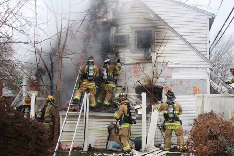 Firefighters in full gear respond to a house fire, climbing ladders and entering a smoke-filled, damaged second-story window. Thick smoke billows from the building as flames are visible and debris surrounds the scene.