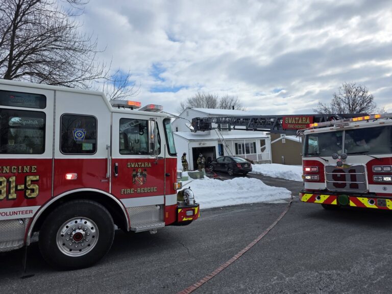 Two red fire trucks are parked outside a house with snow on the ground. Firefighters are near the entrance and a ladder extends from one truck to the roof, under a cloudy sky.