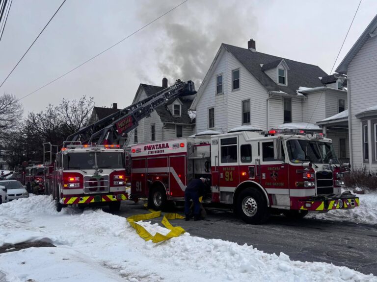 Two fire trucks are parked on a snowy street in front of houses, with firefighters and equipment visible. Smoke rises from one of the houses as a ladder is extended toward the roof.