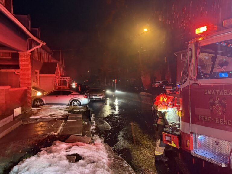 A fire truck with flashing lights is parked on a wet street at night near houses and parked cars; snow and slush cover parts of the sidewalk and street, and streetlights illuminate the scene.