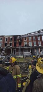 Firefighters in gear stand in front of a row of brick townhouses with visible fire damage and blackened windows. A ladder truck extends to the upper floor as firefighters work on the building.