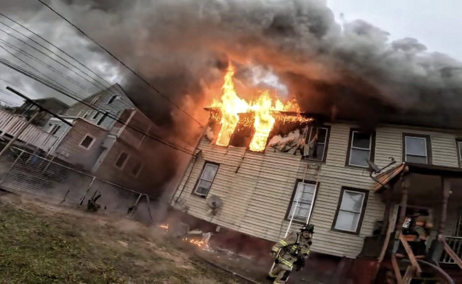 A house with flames and thick smoke billowing from an upstairs window. A firefighter in full gear stands near the burning building, which appears heavily damaged. Power lines cross the scene, and the sky is overcast.