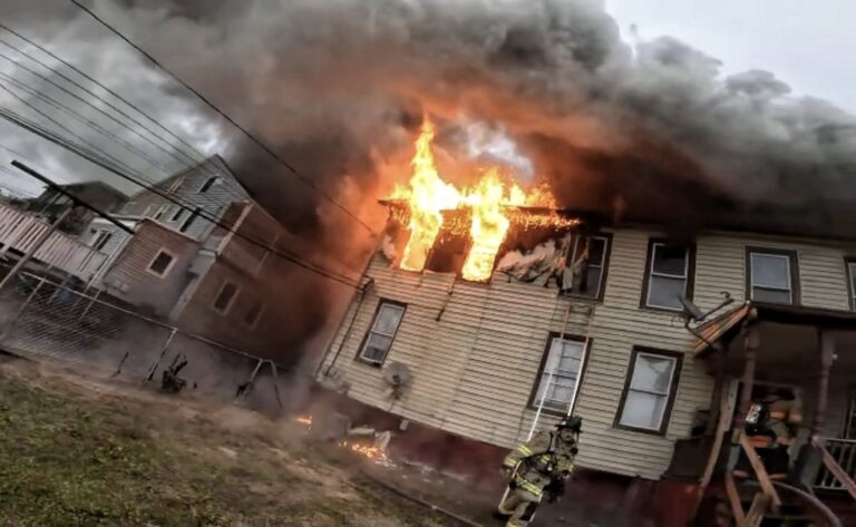 A house with flames and thick smoke billowing from an upstairs window. A firefighter in full gear stands near the burning building, which appears heavily damaged. Power lines cross the scene, and the sky is overcast.
