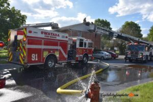 Two fire trucks from Swatara Fire-Rescue spray water on a residential building while firefighters work on the roof; hoses run across wet pavement and water sprays from a fire hydrant in the foreground.